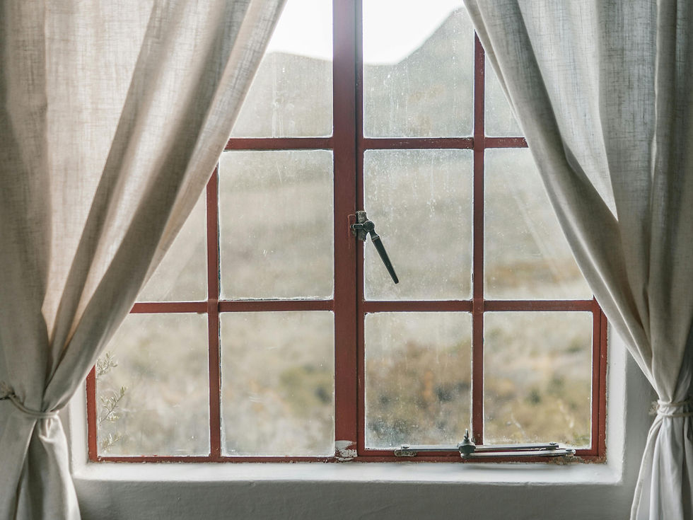 A window with red frame, dusty glass with hard water stains, and beige curtains tied back.
