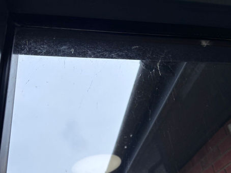 Dirty window with cobwebs and grime. Overcast sky visible outside, with a slight reflection of an interior light and a brick wall edge.