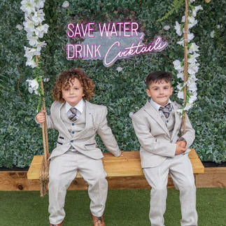 Two young guests in pale 3 piece suits sit on a swing in front of a greenery and flower wall with a neon pink sign