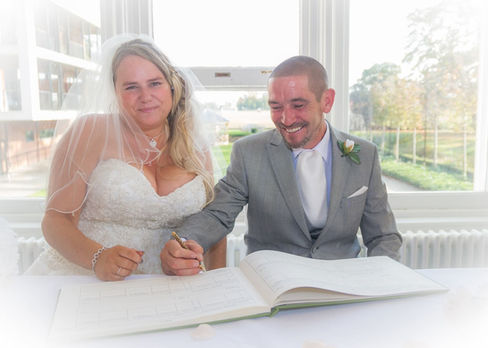 Bride and groom sit at the table to sign the register with the sunlit gardens of Wivenhoe House Hotel behind them