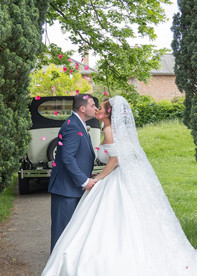 Bride and groom kiss in front of their vintage car transport as petals float down around them