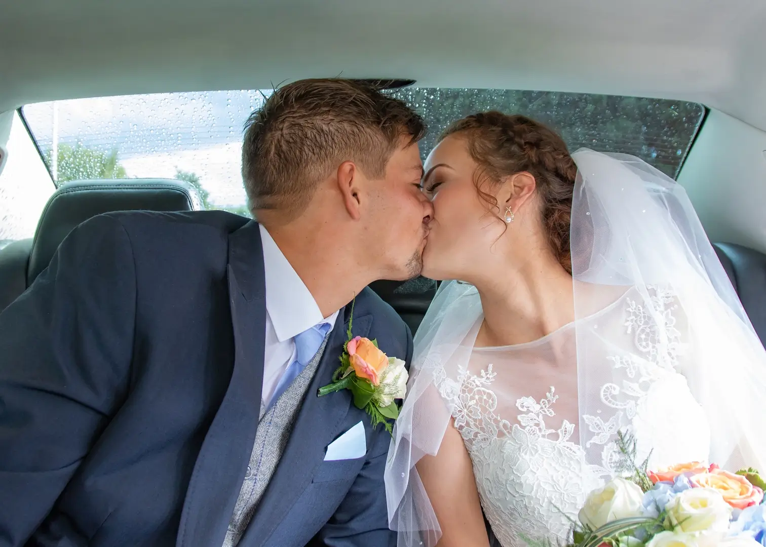 Bride and groom share a kiss in their wedding car as the rain comes down outside