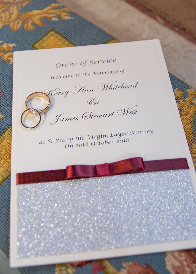 Order of service with burgundy ribbon and silver glitter with the wedding rings lying on top and to one side