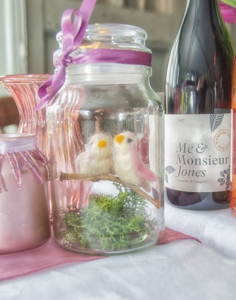 Wedding table decoration made from a glass jar featuring felt songbirds and greenery with a mauve ribbon