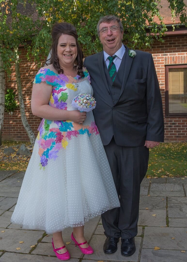 Bride with 1960s hairstyle and white polka dot dress with vibrant colour splotches and vivid pink shoes and a colourful button bouquet stand with her father