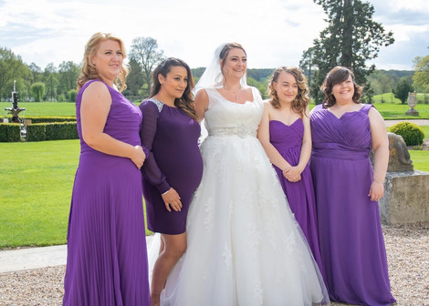 Bride stands on the terrace at Gosfield Hall with her bridesmaids all in cadbury purple dresses