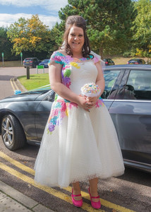 Bride in white dress with harlequin style patches stands by her wedding car