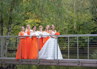 Bride holds a bouqet of orange roses with bridesmaids in orange dresses and white faux fur wraps standing on a bridge