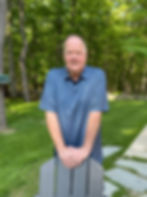 Tommy wearing a blue golf shirt standing behind a chair with trees in the background