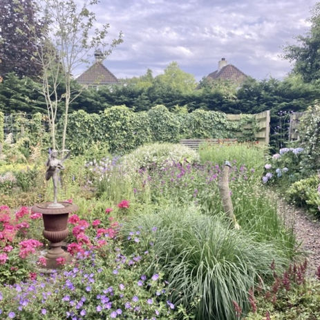 Vanaf het terras is zicht op een tuin vol met kleurrijke beplanting. In deze border staat een beeldje van een vrouw. Een pad loopt naar achteren en rechts van dit pad is ook een plantenborder met kleur.
