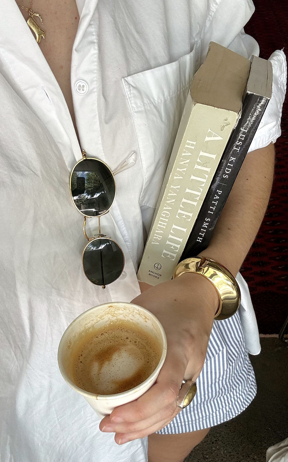 A reader holding books and coffee, wearing a white linen shirt and black and gold sunglasses.
