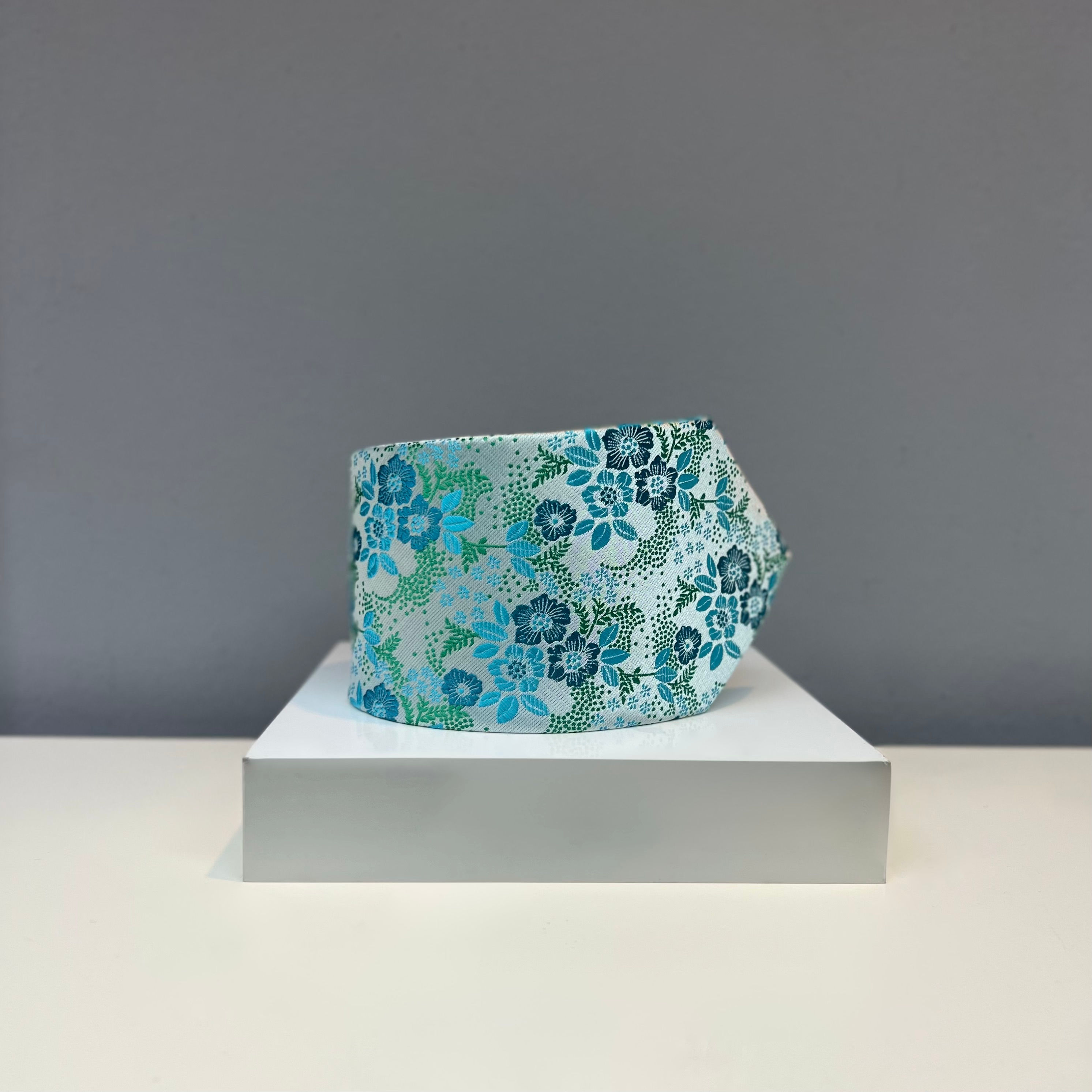 A silver silk tie with teal and aqua floral designs, displayed on a white platform.