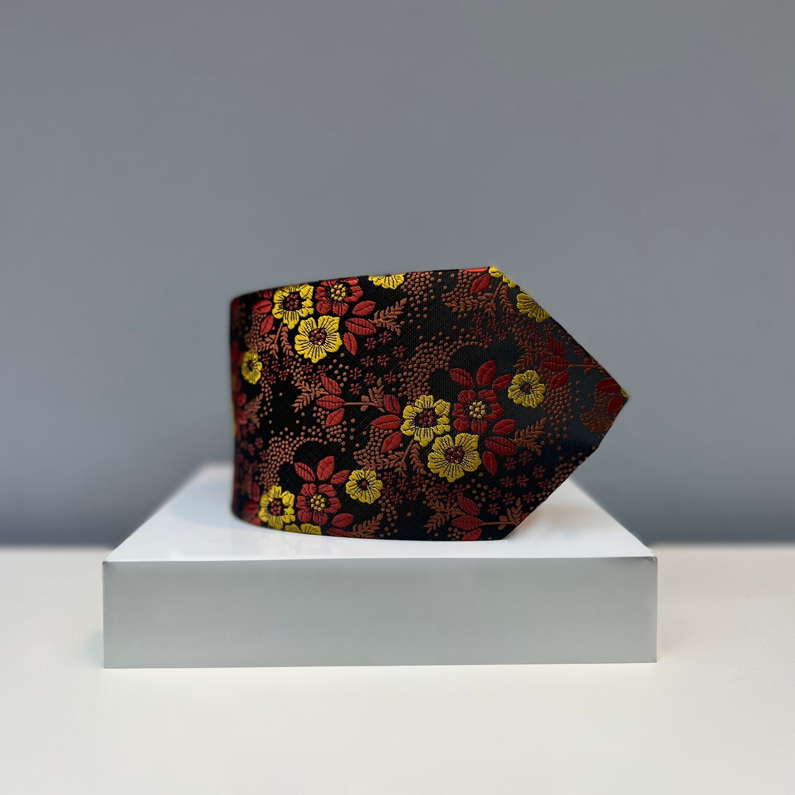 A black silk tie featuring a bright red and yellow floral pattern, displayed on a white platform.