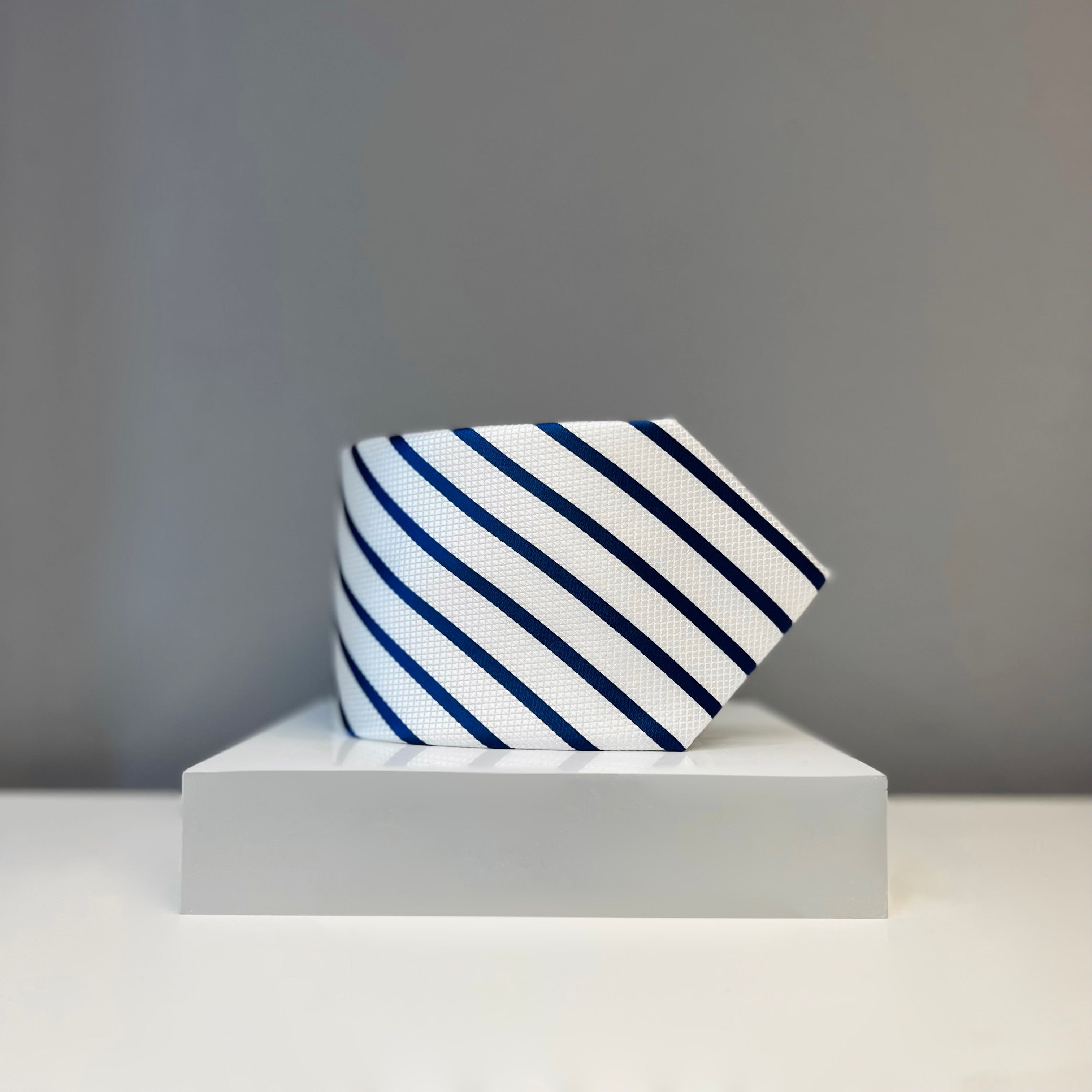 A white silk tie with navy blue diagonal stripes, neatly displayed on a white platform.