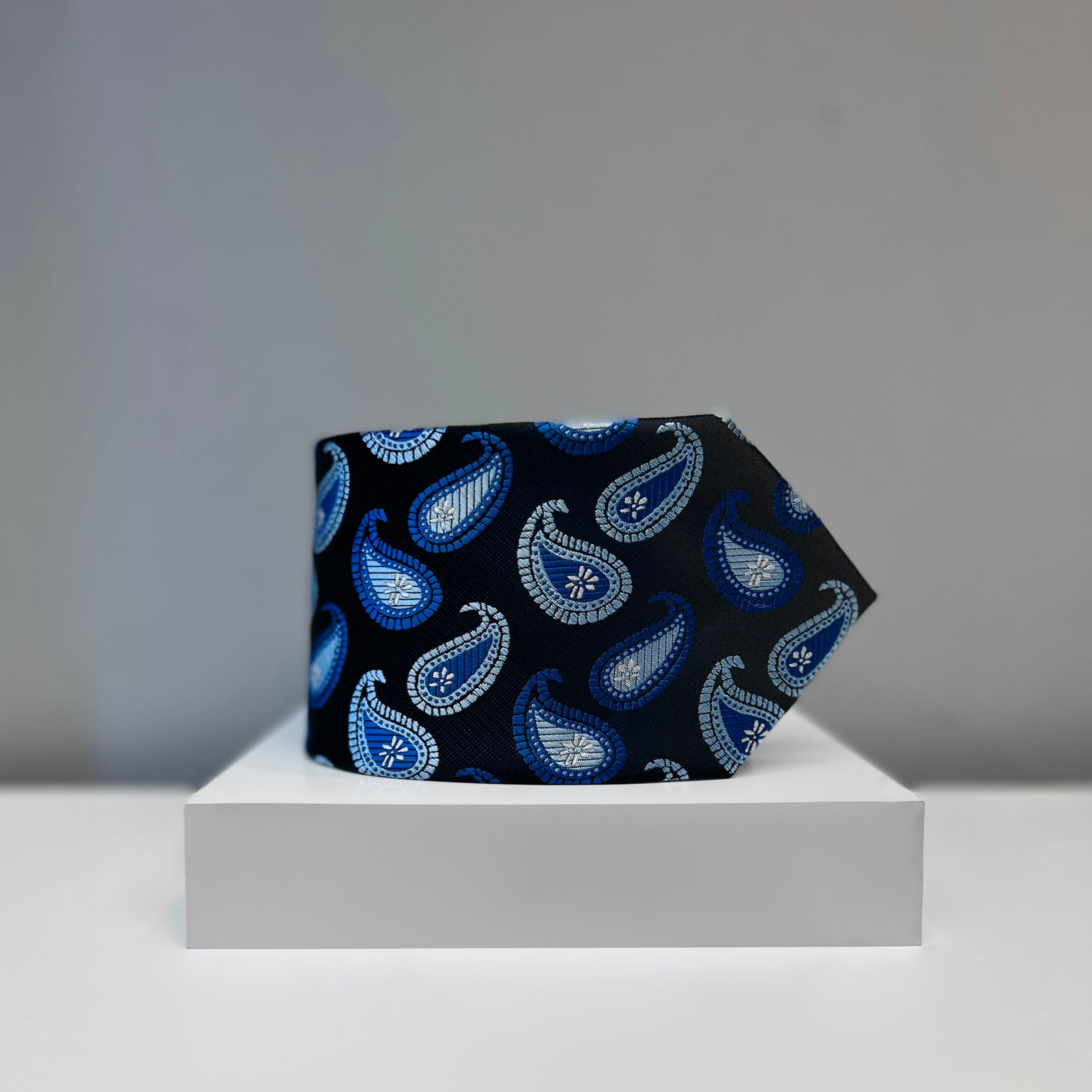 A black silk tie featuring blue and silver paisley patterns, elegantly displayed on a white platform.