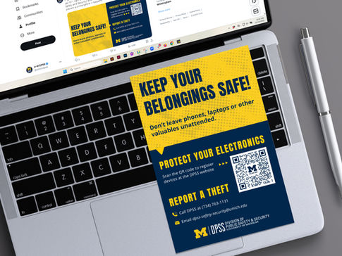 A laptop and pen sitting on a gray surface with a yellow and blue safety flyer resting on the keyboard. The flyer, from the University of Michigan DPSS, advises users to keep their belongings safe and provides instructions for protecting electronics and reporting theft.