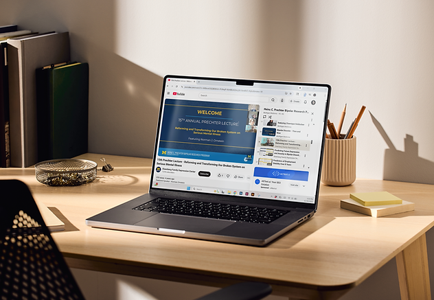 An eye-level, medium shot shows a dark gray laptop open on a light wood desk, angled towards the left. The screen displays a YouTube video titled "15th Prechter Lecture - Reforming and Transforming Our Broken System on Serious Mental Illness."