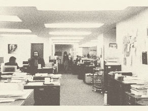 Grainy black-and-white office with people at desks under fluorescent lights, stacks of papers, and a quiet work atmosphere.