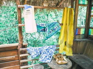 Colorful clothes and scarves hang in a rustic treehouse with jungle views; white shirt reads VISSLA Psychedelic Surfers Society