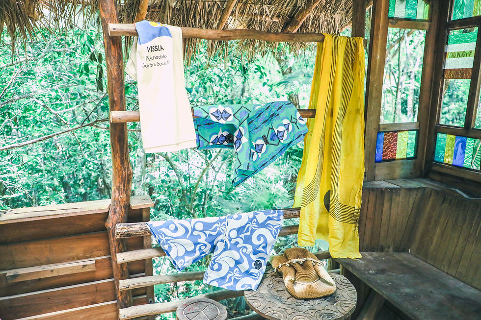Colorful clothes and scarves hang in a rustic treehouse with jungle views; white shirt reads VISSLA Psychedelic Surfers Society