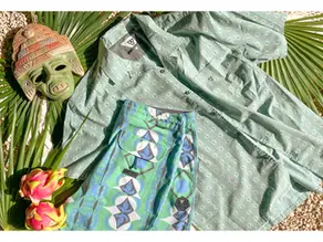 Green patterned shirt and blue-green shorts laid on palm leaves beside a carved mask and dragon fruit on gravel, VISSLA logos visible