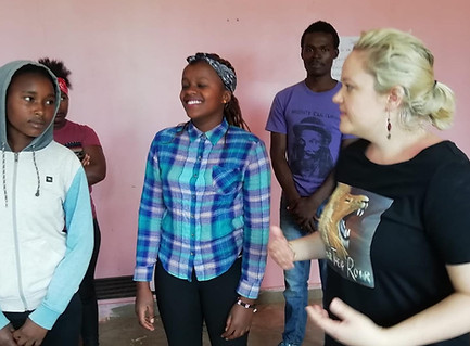 Left to right: Four workshop students, and Clare Wheeler. The student on the far left wears a white and teal hoodie, and looks intently at Clare. Partially obscured, the student next along wears a pink T-shirt with part of a design visible, and a red hair bandana. Their arms are folded. The next student has her eyes closed and is looking slightly to the left, smiling with a broad grin. She is wearing a plaid button up shirt, in teal, dark blue, and white. She has a black and white hair bandana. The next student looks at the camera with his hands together. He is wearing a purple T-shirt with an image of a beared man with long hair on it, and jeans. Clare is looking to the left and gesturing with her hands in front of her, palms upwards. She has her hair tied back, is wearing simple drop earrings and has a T-shirt with a lion roaring on it. Part of the text can be made out: 'Roar'. They stand in a room with pink walls.