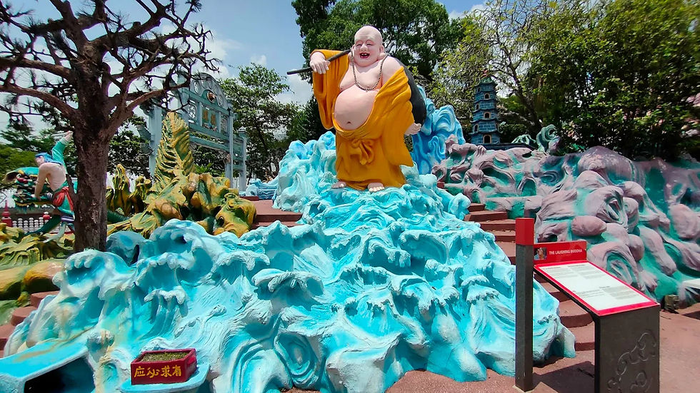 Laughing Buddha statue in yellow robes stands on blue waves in a park. Trees and colorful sculptures surround, sign reads "The Laughing Buddha".