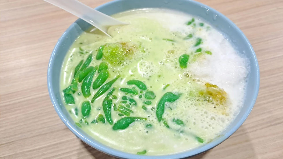 A blue bowl of green cendol dessert with coconut milk and ice on a wooden table. A plastic spoon rests inside, creating a refreshing mood.