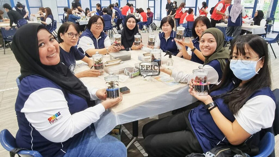 Group of women smiling and holding terrariums at a workshop table. They wear navy shirts with "EPIC Workshops" sign visible. Indoor setting.