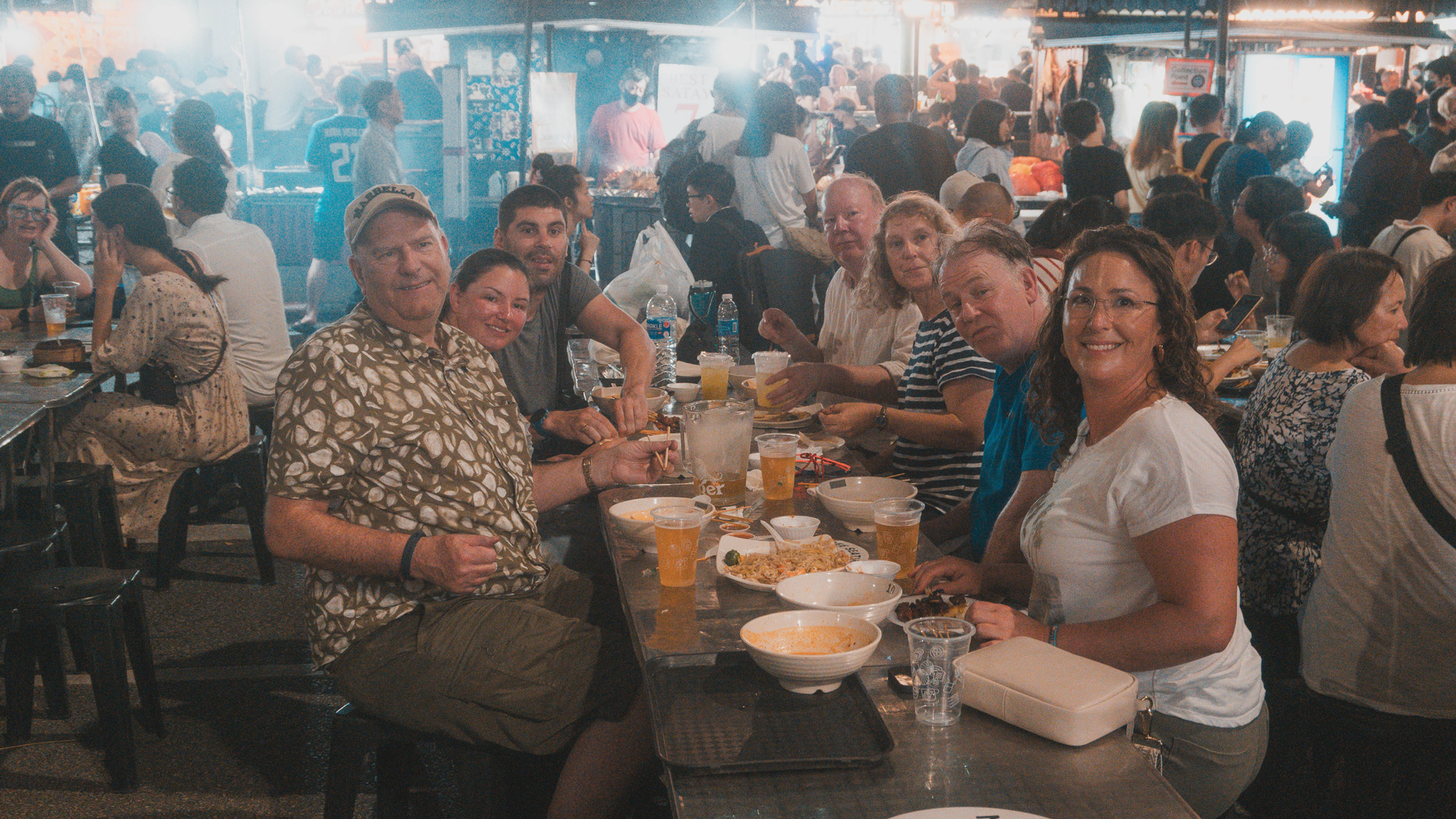 Smiling group at a crowded night market food stall, eating and drinking at a table under bright lights; signs read SAINY 7 and SATAY 8