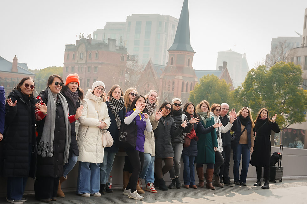Group of smiling people in winter coats wave on a city sidewalk, with a church spire and buildings in the hazy background.