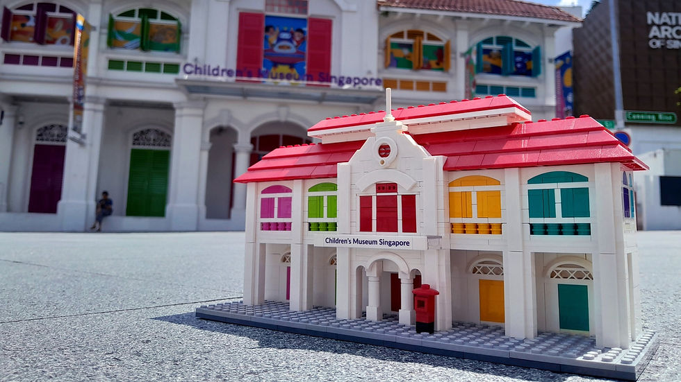 LEGO model of Children's Museum Singapore with colorful windows and a red roof sits in front of the real building. Bright, sunny day.