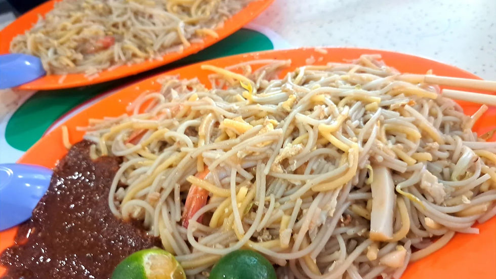 Orange plates with noodles, prawns, cuttlefish, sambal, and lime slices. Blue utensils on a speckled surface create a casual dining scene.