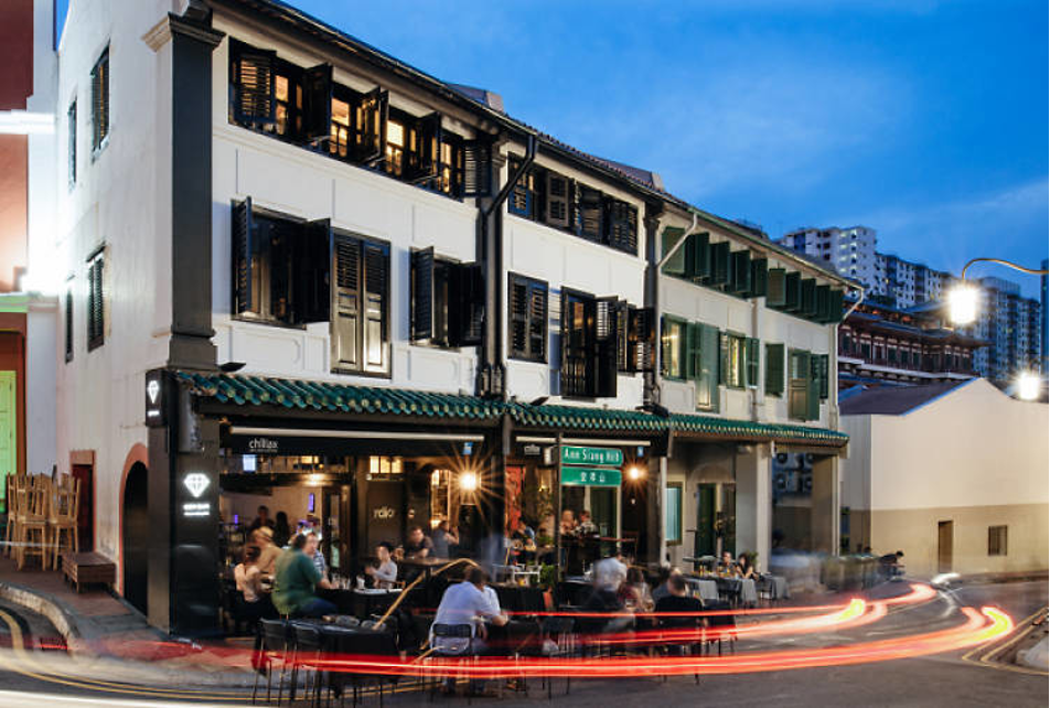 People dine outside a corner restaurant named Ann Siang Hill at dusk. Motion blur of passing cars. Historic building, blue sky.