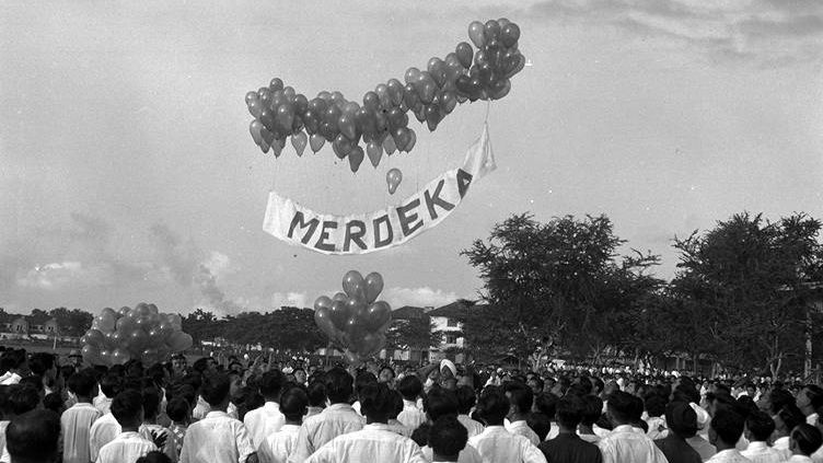 Crowd watches balloons lifting a "Merdeka" banner skyward. Trees and buildings in the background create a festive, hopeful atmosphere.