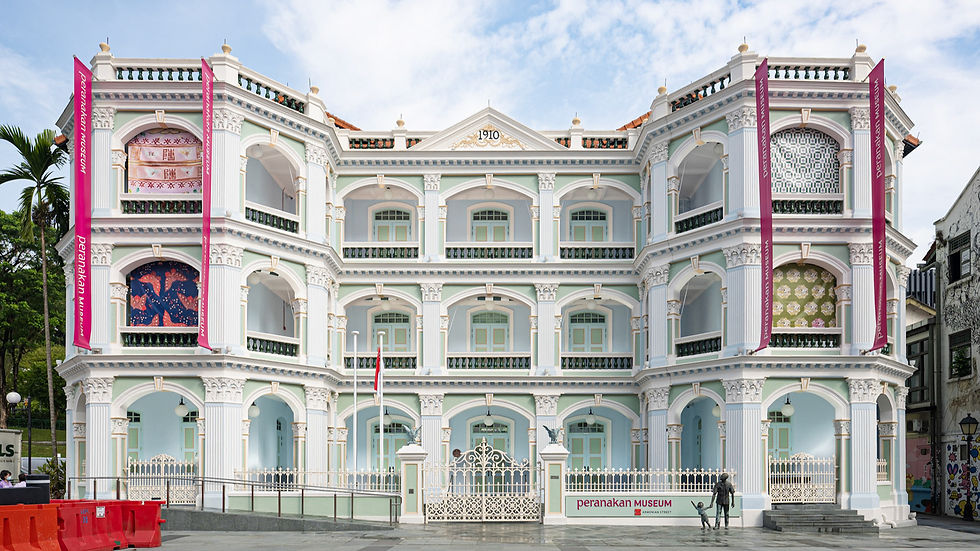Ornate white building with pastel green trim, labeled "Peranakan Museum." Pink banners and intricate patterns adorn it. Bright, sunny day.
