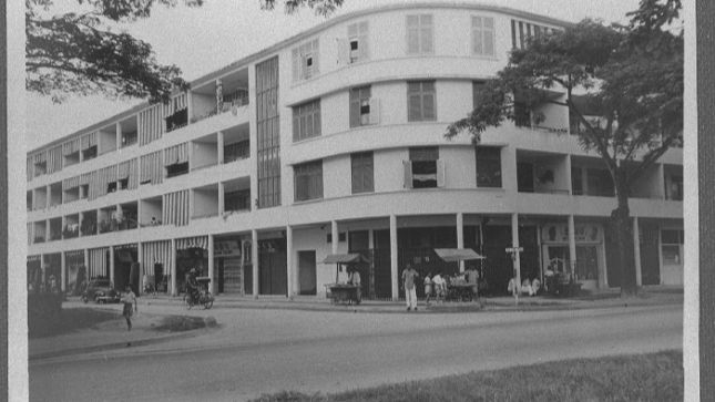 Historic black-and-white photo of a curved, multi-story building with people and bicycles on the sidewalk. Trees frame the scene.