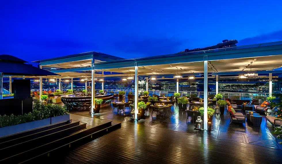 Rooftop bar with tables and lush plants under a blue evening sky. The setting is elegant, well-lit, and overlooks a city skyline.