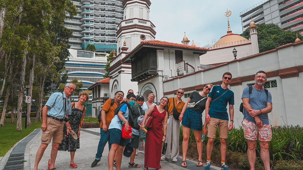 Group of smiling people pose in front of an ornate mosque with a gold dome and tall buildings in the background. Lush trees add greenery.