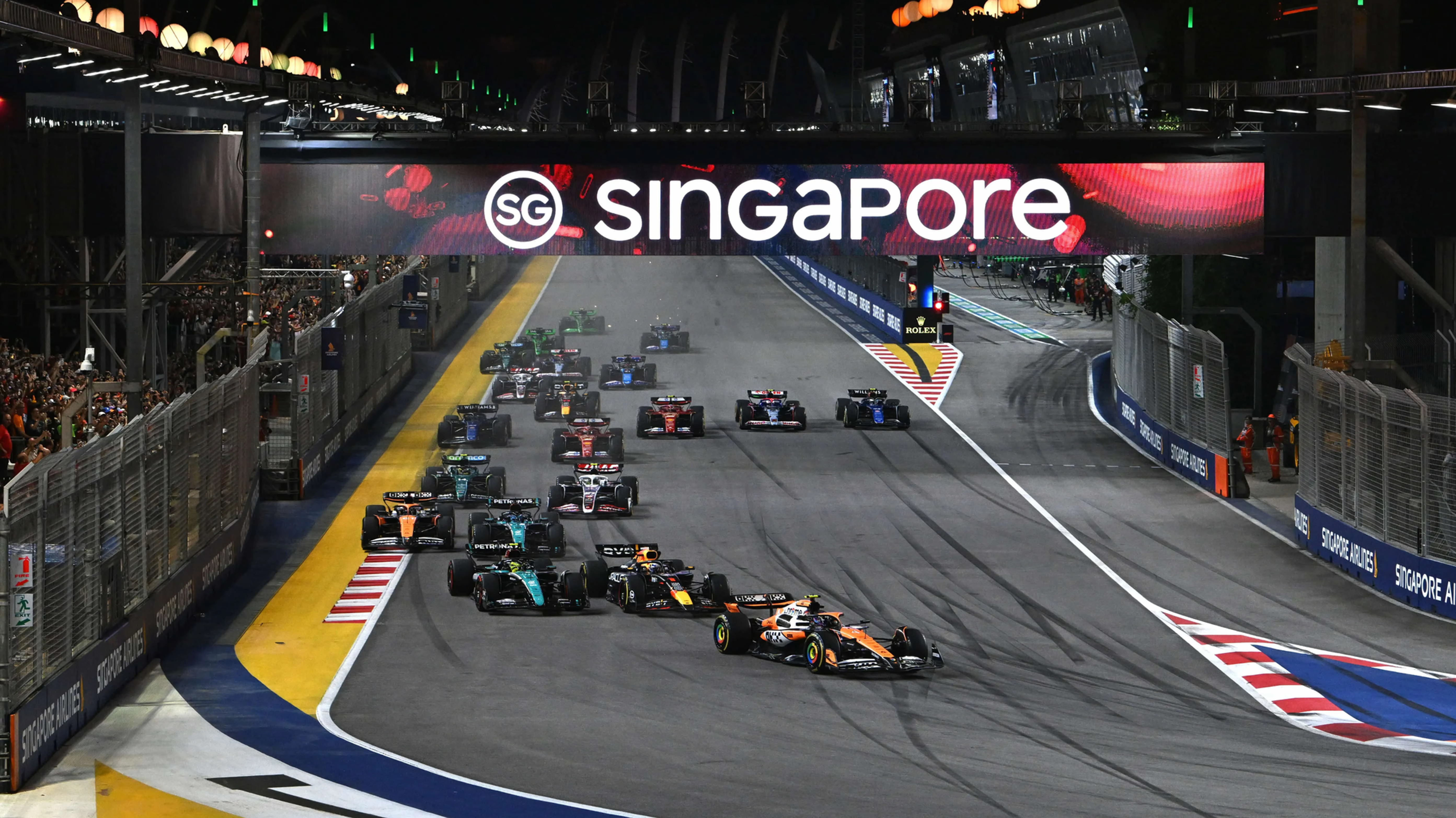 Formula 1 cars race through Singapore’s night circuit under a glowing SG Singapore banner, with crowds behind fencing.