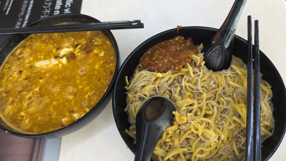 Two bowls of food; one with spicy orange soup, the other with yellow noodles topped with chili paste. Black chopsticks and spoons included.