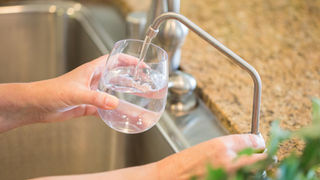 Hands filling a glass with water from a sleek metal faucet. Background shows a granite countertop and a sink, with a hint of greenery.