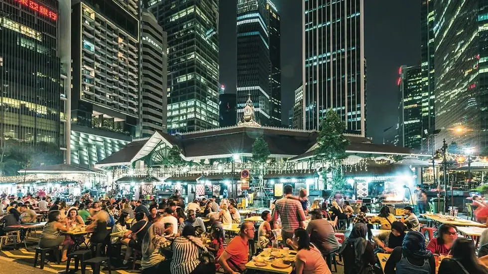 Nighttime street market with crowds dining at tables, surrounded by tall skyscrapers. Bright lights and lively atmosphere.