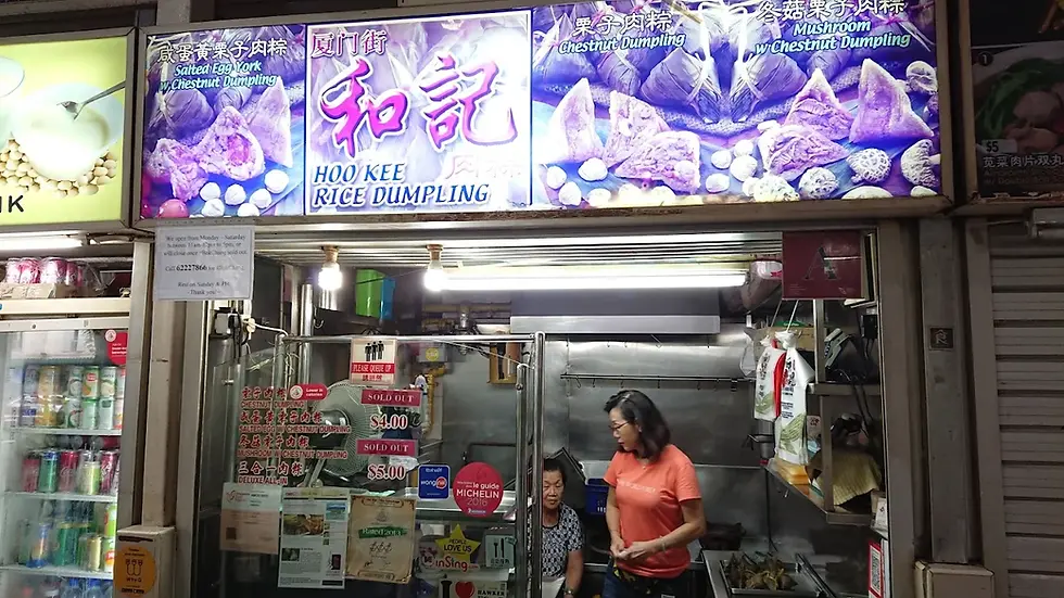 Two women in front of Hoo Kee Rice Dumpling stall. The sign features bright purple images of dumplings. Text shows menu and sold-out items.