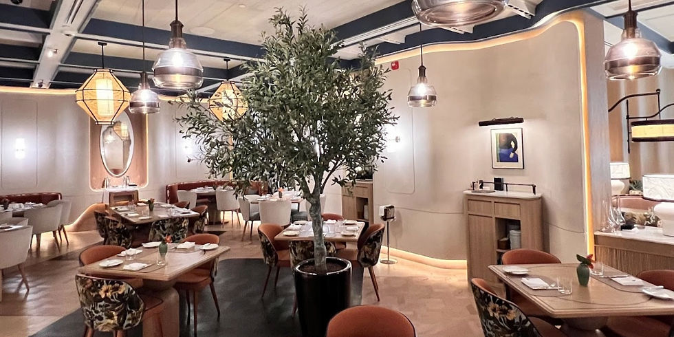 Elegant restaurant with cozy, modern decor, featuring a central potted tree, warm lighting, art on walls, and set tables with floral chairs.