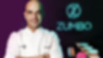 Bald man in a white chef's coat smiles beside neon "ZUMBO" sign. Blue and pink macarons on table. Dark brick backdrop.