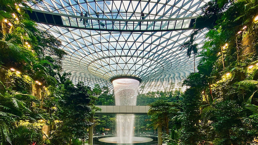Tropical indoor garden with lush greenery, a central waterfall, and a glass-domed ceiling. Walkway above with silhouetted people.