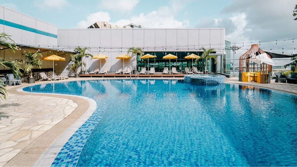 Rooftop pool with clear blue water, yellow umbrellas, palm trees, and lounge chairs. Bright, sunny day with a modern building backdrop.