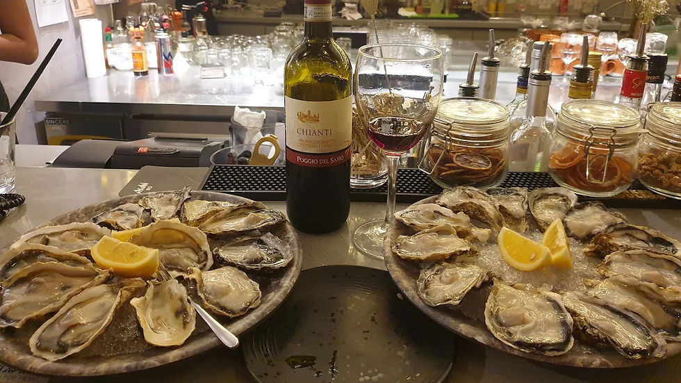 Oysters with lemon slices on plates, Chianti wine bottle, and glass on bar counter. Background has jars, glasses, and drink bottles.
