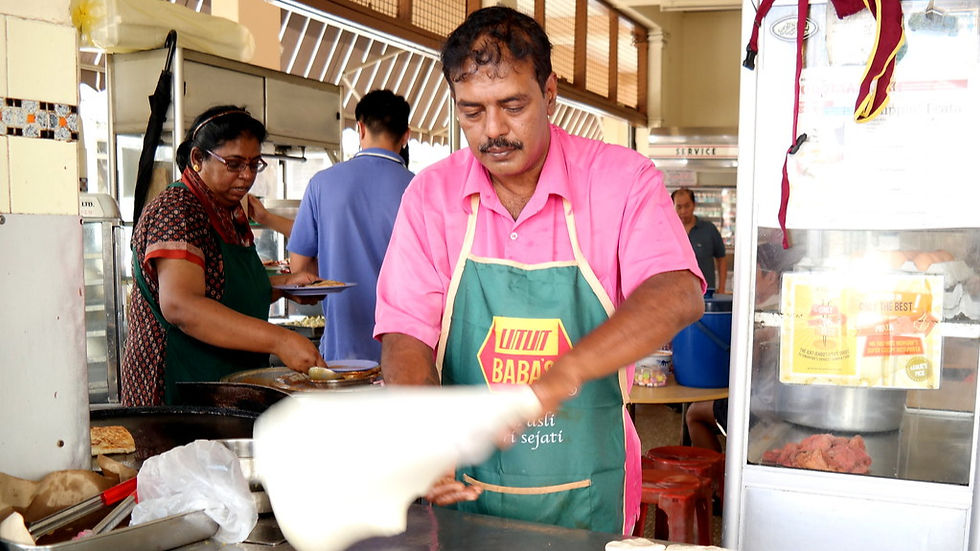 Man in pink shirt making food with woman assisting in market stall. Wearing green apron with red and yellow text. Busy atmosphere.